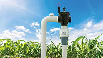 Close-up of a Senninger® PRU-LV Limit Valve installed on a field irrigation system, with healthy corn plants and a bright blue sky in the background.