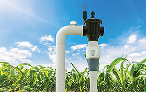 Primer plano de una válvula limitadora Senninger® PRU-LV instalada en un sistema de riego de campo, con plantas de maíz sanas y un cielo azul brillante de fondo.