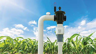 Close-up of a Senninger® PRU-LV Limit Valve installed on a field irrigation system, with healthy corn plants and a bright blue sky in the background.